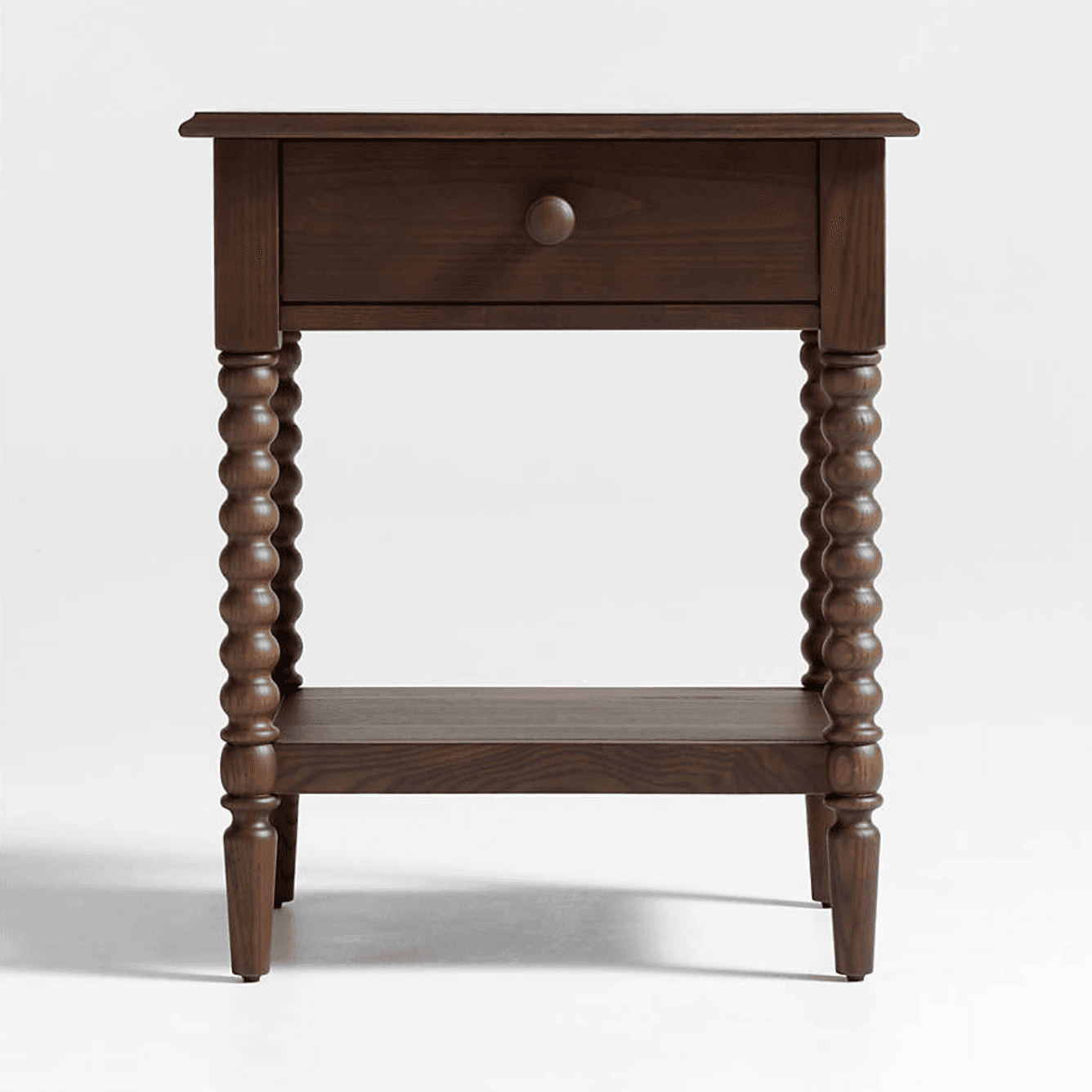 A dark brown wooden nightstand featuring a single top drawer with a round wooden pull knob and an open lower storage shelf. The table is supported by four distinctive bobbin-turned legs, which taper slightly at the bottom. The wood exhibits a visible grain pattern, suggesting a solid wood construction with a rich, dark stain. — 3D model