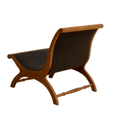 A low-profile X-frame lounge chair constructed from richly toned wood and upholstered in dark material. The frame features curved, intersecting legs that form an 'X' shape, connected by a turned stretcher bar at the back. The seat and backrest are simple, suspended panels of dark upholstery, providing a comfortable recline. The wooden frame has smooth, rounded edges, indicative of quality craftsmanship. — 3D model