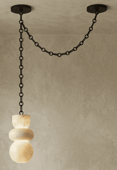 A contemporary pendant light featuring a sculptural shade crafted from translucent alabaster, suspended by a black metal chain system. The shade is composed of three stacked, organic, rounded forms, illuminated from within to highlight the natural veining and luminous quality of the stone. The suspension system consists of a long chain dropping from one ceiling mount, connecting to the light fixture, and a shorter chain extending horizontally to a second ceiling mount, creating a distinctive draped effect. — 3D model