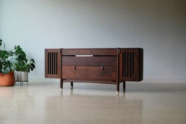 A long, low, dark wooden cabinet with a distinct mid-century modern design. It features slatted sections on the curved ends, a central open shelf, and multiple drawers and cabinet doors below, all adorned with small brass pull handles. The cabinet is supported by angled, tapered legs with brass ferrules at the bottom. — 3D model