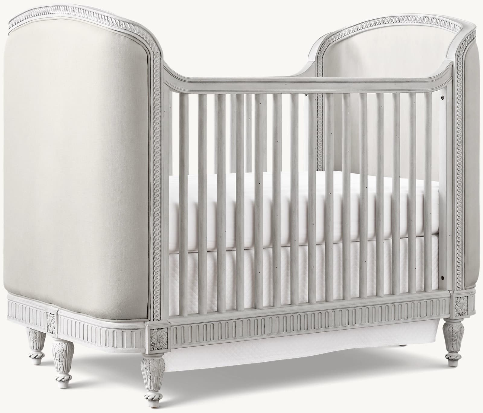 A classic-style crib with upholstered side panels and a slatted front and back. The frame is painted a light, neutral color and features decorative molding and carved details on the legs and rails. It includes a mattress with white bedding. — 3D model