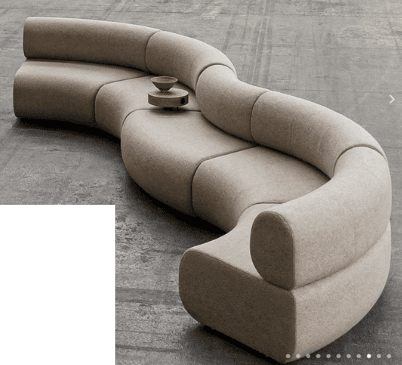 A modular, serpentine-shaped sofa with a low profile. The sofa is upholstered in a textured, light-colored fabric and consists of multiple interlocking curved sections, creating a flowing, organic form. It appears to be designed to accommodate multiple seating arrangements and potentially a central element like the small wooden table with a bowl, which is positioned on one of the seat sections. — 3D model