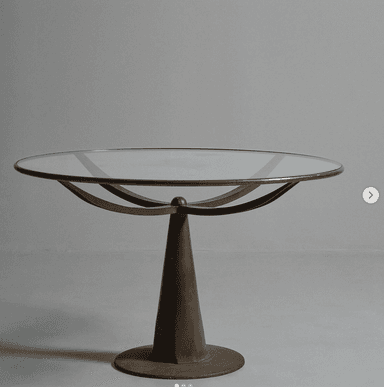A round accent table with a glass top and a metal pedestal base. The base has a conical shape that flares out to a circular foot. Underneath the glass top, there are two curved metal supports that cross each other. — 3D model