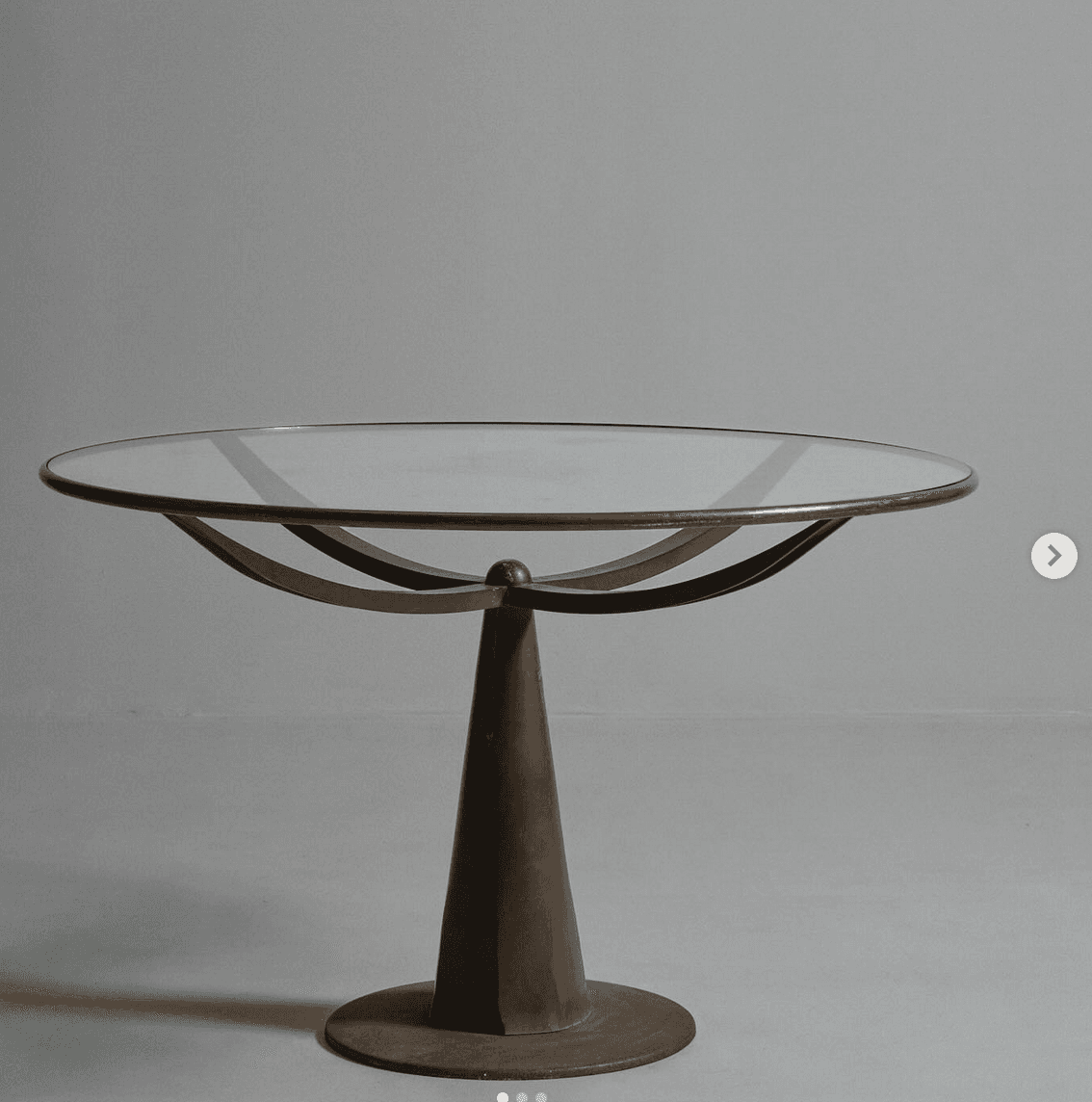 A round accent table with a glass top and a metal pedestal base. The base has a conical shape that flares out to a circular foot. Underneath the glass top, there are two curved metal supports that cross each other. — 3D model
