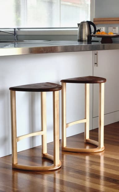 A wooden bar stool with a triangular, slightly dished seat and three cylindrical legs. One leg is positioned at the front and two at the back. A curved stretcher connects the two back legs near the bottom, and a diagonal stretcher connects the front leg to the rear left leg. The stool is constructed from light-colored wood with a visible grain. — 3D model