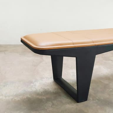 A long, rectangular bench featuring a padded, tan upholstered seat with stitched detailing, supported by a minimalist black metal frame with inverted V-shaped legs. — 3D model