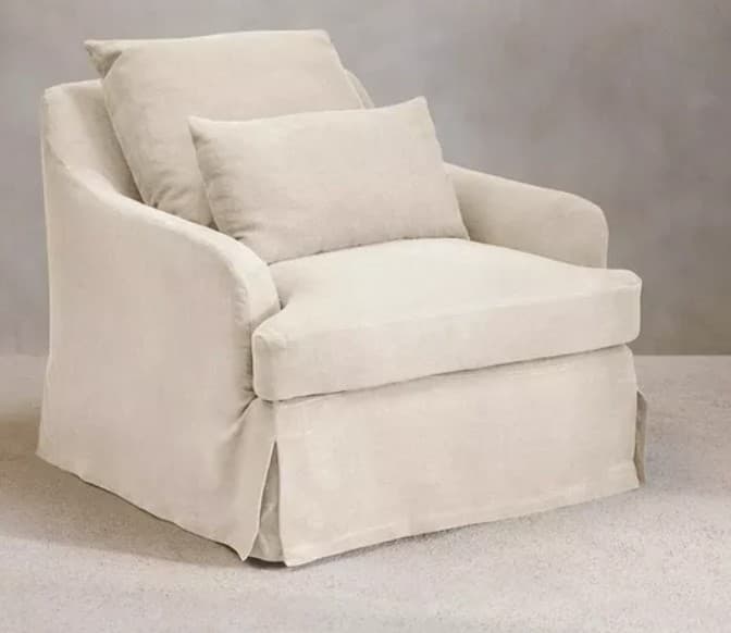 A light-colored, upholstered armchair featuring a loose slipcover design and a skirted base. It has wide, slightly rolled armrests and includes two loose, rectangular cushions for the seat and back, plus a smaller lumbar or accent cushion. The overall aesthetic is casual and comfortable. — 3D model