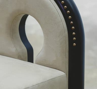 A contemporary chair with a distinctive U-shaped backrest that forms a cutout. The seat and the curved portion of the backrest are upholstered in a light beige, suede-like fabric. The frame is composed of dark, possibly black or dark wood, elements. The outer curved frame of the backrest and the vertical supports of the legs are adorned with a series of small, brass or gold-colored studs. The legs are thick and rectangular, with a dark finish. The chair's design is modern and somewhat sculptural, featuring a negative space in the upper backrest. — 3D model