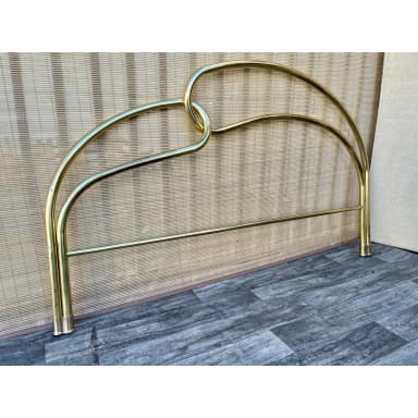 A polished metal headboard with a curved design. The headboard features two parallel horizontal bars supporting an arched top section. The arched section is formed by two tubular elements that loop and interlock in the center, creating a symmetrical, modern aesthetic. The vertical supports at each end are cylindrical, with visible fasteners or adjustment points at the base. — 3D model