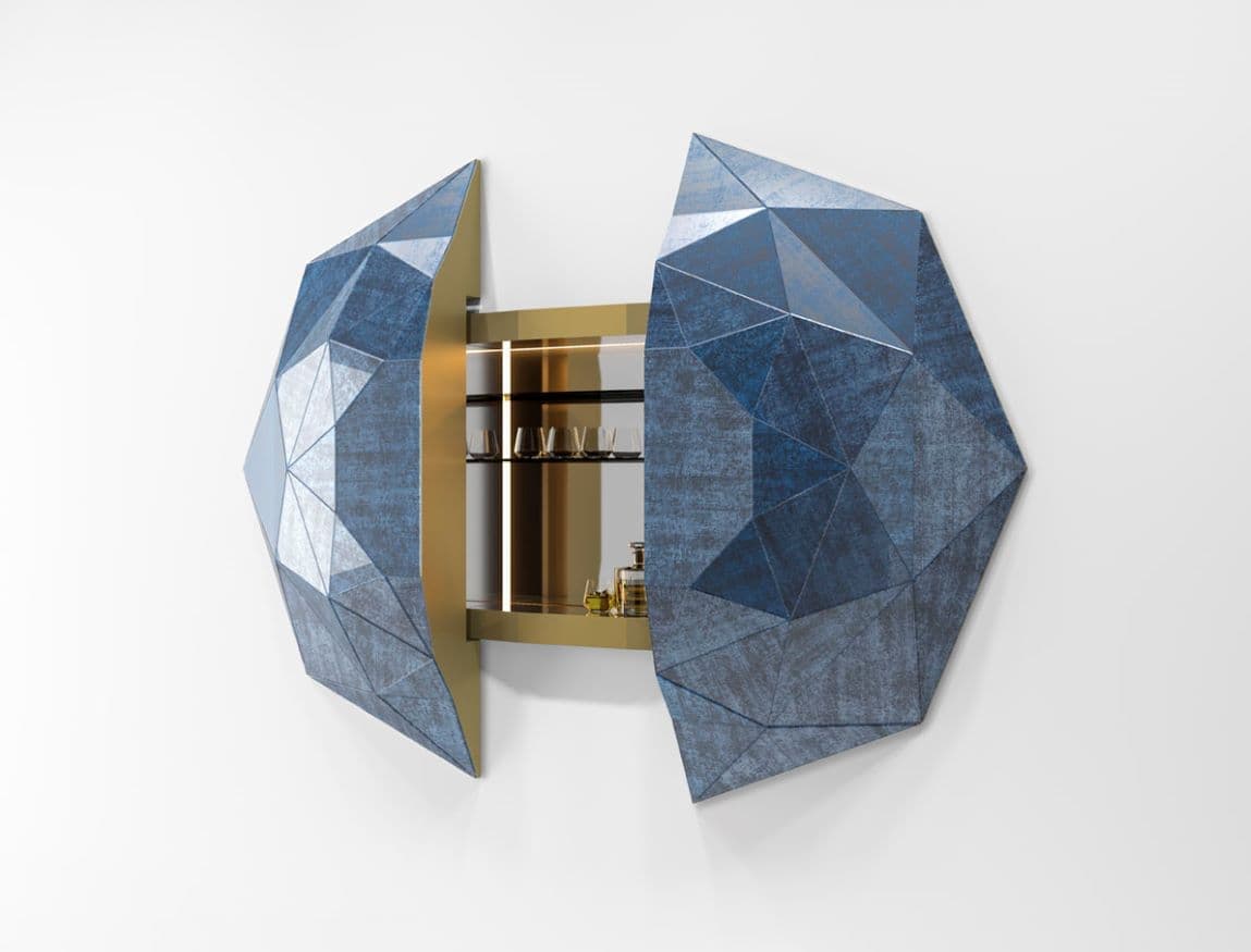 A striking wall-mounted bar cabinet featuring a highly geometric and faceted exterior, resembling a gemstone or crystal formation. The exterior panels have a textured, distressed blue finish, while the interior reveals a luxurious polished brass or gold-toned metal with integrated LED lighting. The cabinet opens in two halves, revealing shelves designed to hold barware and bottles. The overall design is contemporary and sculptural. — 3D model