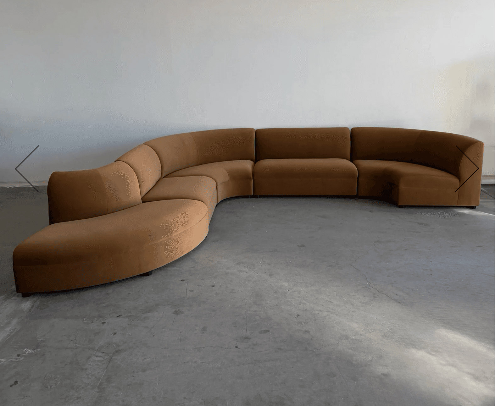 A large, modular, curved sectional sofa with a low profile and rounded edges. It appears to be upholstered in a soft, velvety fabric. The sections are arranged in a crescent shape, with one end featuring a chaise-like extension and the other a more traditional closed end. — 3D model