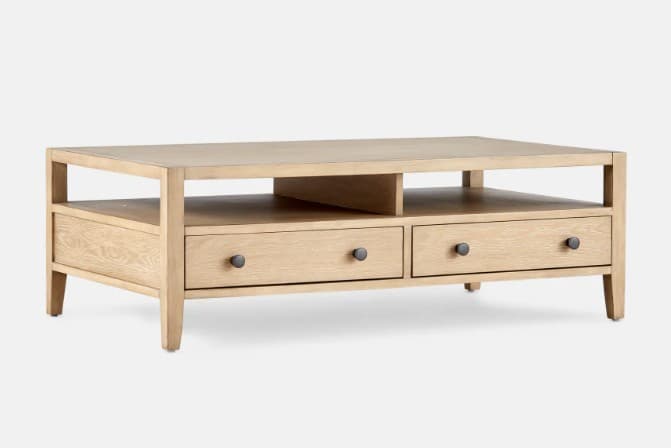 A light-colored wooden coffee table with a rectangular top and an open lower shelf. It features three drawers with dark, circular knobs. The overall design is minimalist with clean lines and tapered legs. — 3D model