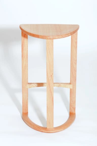 A wooden bar stool with a triangular, slightly dished seat and three cylindrical legs. One leg is positioned at the front and two at the back. A curved stretcher connects the two back legs near the bottom, and a diagonal stretcher connects the front leg to the rear left leg. The stool is constructed from light-colored wood with a visible grain. — 3D model