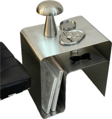 A modern, metallic end table with a minimalist, curved design. It features a continuous surface that forms the top and sides, with an open compartment beneath for storage. The table has a secondary lower shelf that is partially visible and appears to have a cutout design. It is suitable for placement next to a sofa or bed. — 3D model