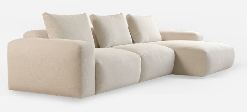 A contemporary, plush, light beige sectional sofa featuring a modular design with clean lines and a low profile. It consists of multiple sections, including a three-seat segment and an extended chaise lounge, all upholstered in a textured fabric. The backrests are composed of loose, rectangular cushions, complementing the soft, rounded appearance of the base and arms. — 3D model