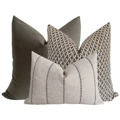 A set of three decorative throw pillows. One pillow is a solid dark grey, likely velvet or a similar soft fabric. Another pillow features a repeating pattern of dark brown stylized leaf motifs on a cream background. The third pillow is rectangular and oriented horizontally, with a textured cream fabric and vertical stripes composed of dashed lines in a dark grey or brown hue. The pillows are arranged in a layered composition. — 3D model