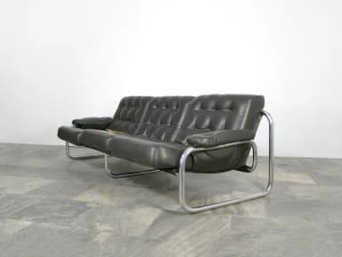 A three-seater sofa featuring black button-tufted leather upholstery and a polished chrome tubular steel frame. The cushions are generously padded, and the armrests are integrated into the frame structure with matching leather padding. The design suggests a mid-century modern aesthetic, emphasizing both comfort and a sleek profile. — 3D model