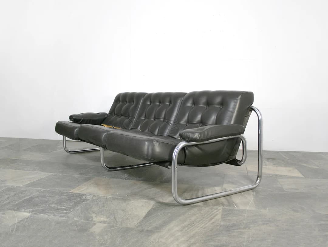 A three-seater sofa featuring black button-tufted leather upholstery and a polished chrome tubular steel frame. The cushions are generously padded, and the armrests are integrated into the frame structure with matching leather padding. The design suggests a mid-century modern aesthetic, emphasizing both comfort and a sleek profile. — 3D model