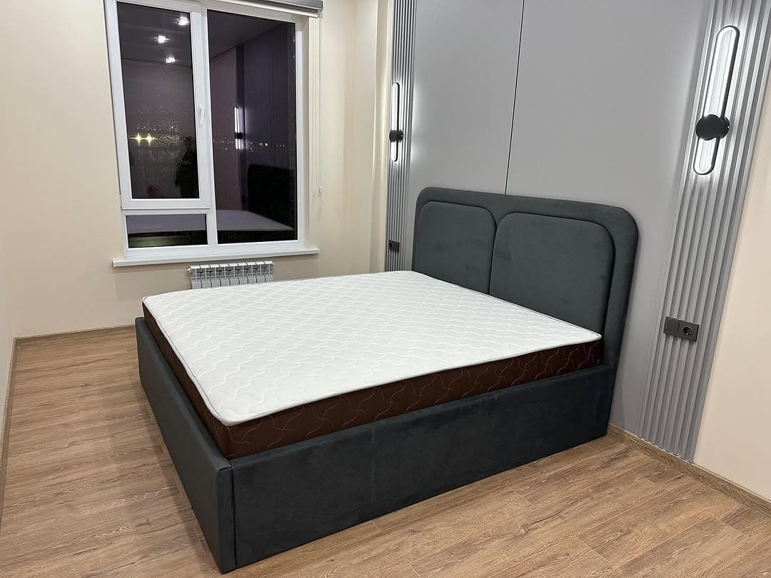 A contemporary upholstered platform bed featuring a dark gray fabric frame and a matching headboard. The headboard is designed with two subtly rounded, padded panels. A white mattress with a patterned top surface and brown sides rests on the platform, indicating it is likely a new or well-maintained item. The bed is situated on a light wooden floor in a room with light-colored walls and modern wall lighting fixtures. — 3D model