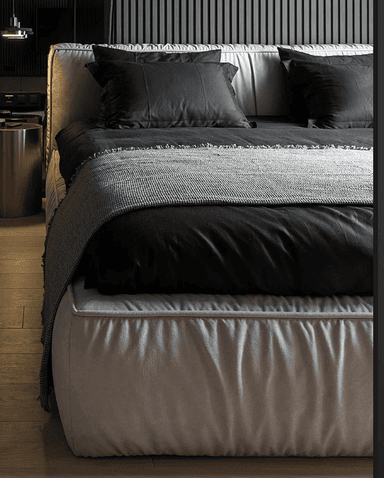 A modern upholstered platform bed featuring a low-profile base and a tall, pleated headboard. The bed is covered in a light beige or off-white fabric, with a distinctive vertical pleated texture on both the headboard and the perimeter of the bed base. It is dressed with black bedding and a grey knitted throw blanket. — 3D model