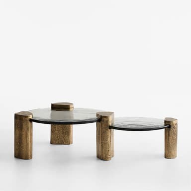 A contemporary coffee table with a circular, smoked glass top supported by four uniquely shaped, textured metallic legs. The legs appear to be cylindrical with irregular, rough surfaces and are finished in a bronze-like hue. The overall design is minimalist yet impactful, with the textured legs contrasting with the smooth glass. — 3D model
