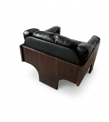 A robust lounge chair featuring a distinctive dark wood veneer frame, likely rosewood, with deep-set black leather cushions. The chair's base forms a solid, geometric structure with cut-out arches, providing a foundational aesthetic. The upholstery consists of plush, padded black leather, including a seat cushion, backrest, and armrests, suggesting a comfortable seating experience. The overall design exhibits characteristics of Mid-Century Modern style with a touch of architectural solidity. — 3D model