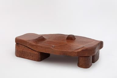 A unique, low-profile coffee table carved from wood. The tabletop features an organic, undulating surface with a textured finish, resembling natural erosion or flowing water. It is supported by four block-like legs, also made of wood. A single, small rectangular block rises from the center of the tabletop. The overall aesthetic is rustic and sculptural, with visible tool marks and a rich wood grain. — 3D model