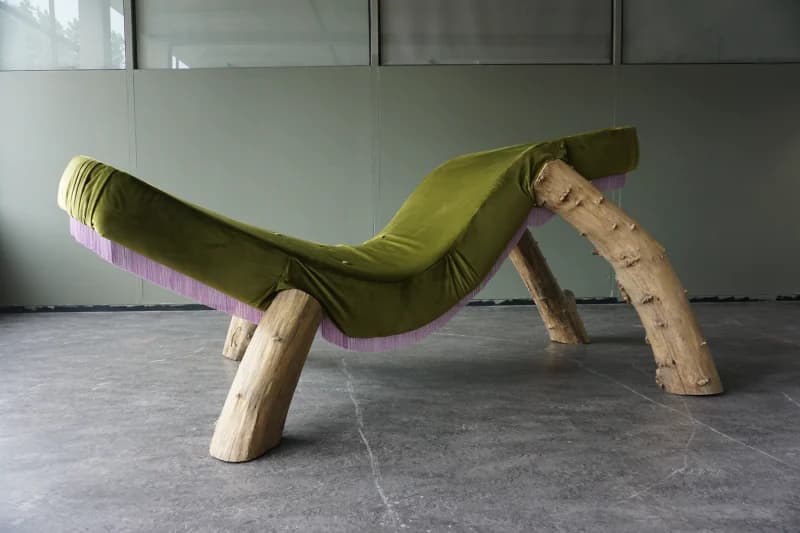 A chaise lounge with a distinctive organic design. The seating surface is upholstered in a plush, olive-green velvet fabric with subtle pleating. It is supported by two pairs of thick, natural wood logs that serve as legs, retaining their bark and irregular shapes. A band of light purple fringe runs along the underside edge of the upholstered seat. — 3D model