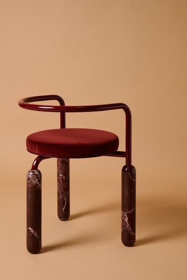 A contemporary accent chair featuring a circular deep red velvet upholstered seat, supported by a curved, glossy dark red metal frame that also forms the backrest and partial armrests. The chair stands on three robust, cylindrical legs made of polished dark red marble with white veining. — 3D model