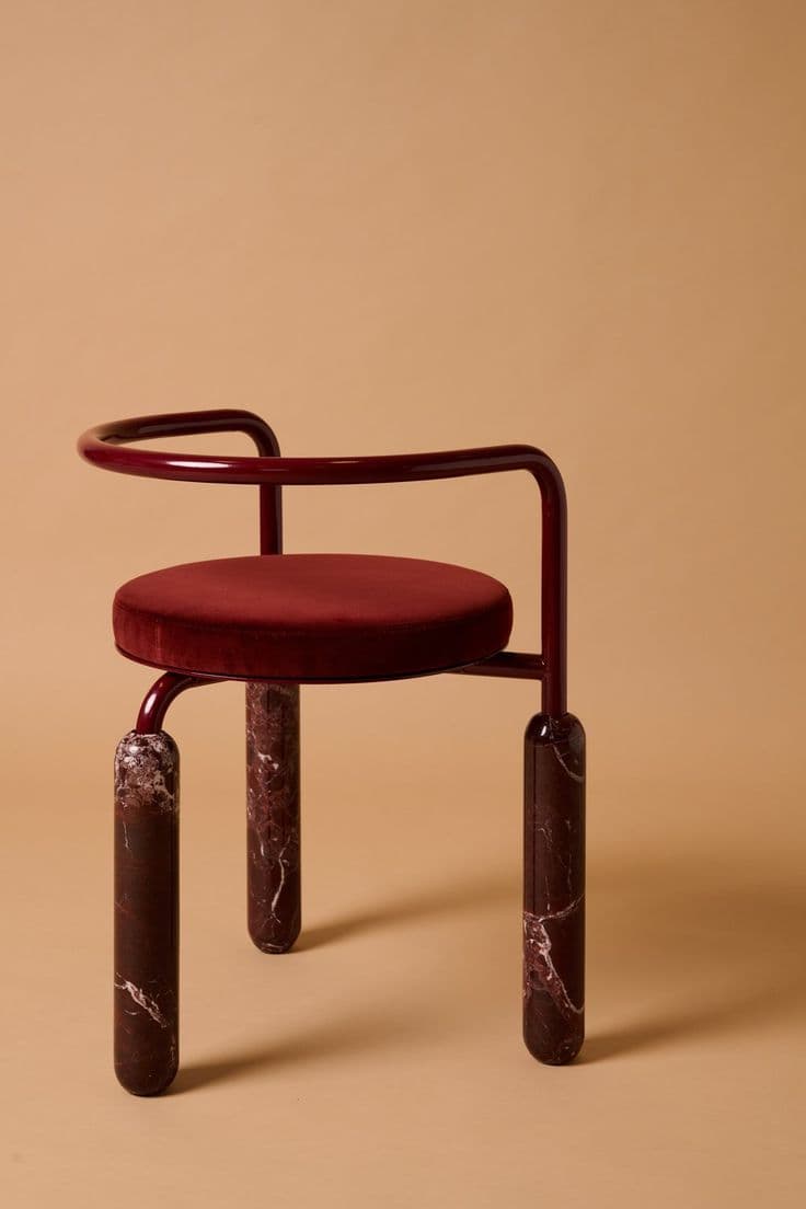 A contemporary accent chair featuring a circular deep red velvet upholstered seat, supported by a curved, glossy dark red metal frame that also forms the backrest and partial armrests. The chair stands on three robust, cylindrical legs made of polished dark red marble with white veining. — 3D model
