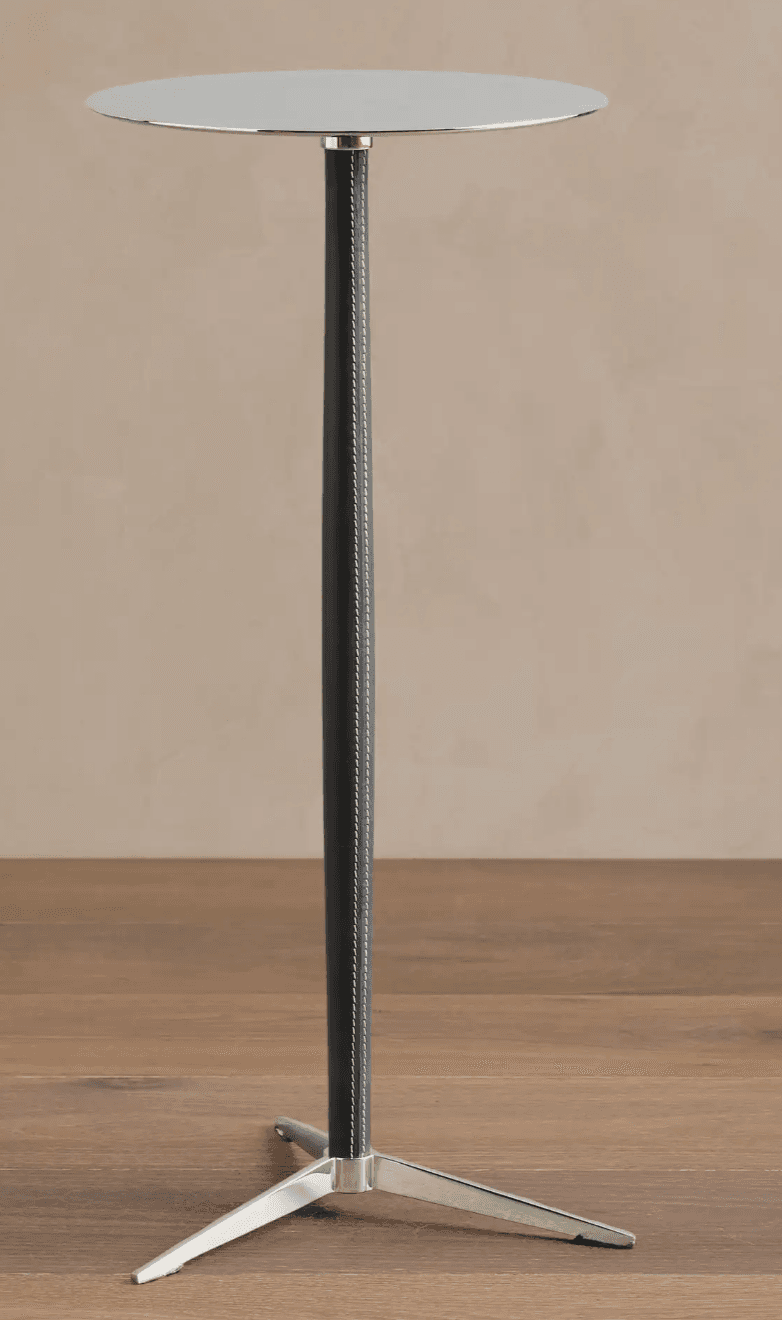 A tall, slender side table with a circular, reflective top. The central column is covered in dark material, possibly leather, with prominent white stitching along its length. The base is composed of three metallic legs splayed outwards, providing stability. The overall aesthetic suggests a modern or contemporary design. — 3D model