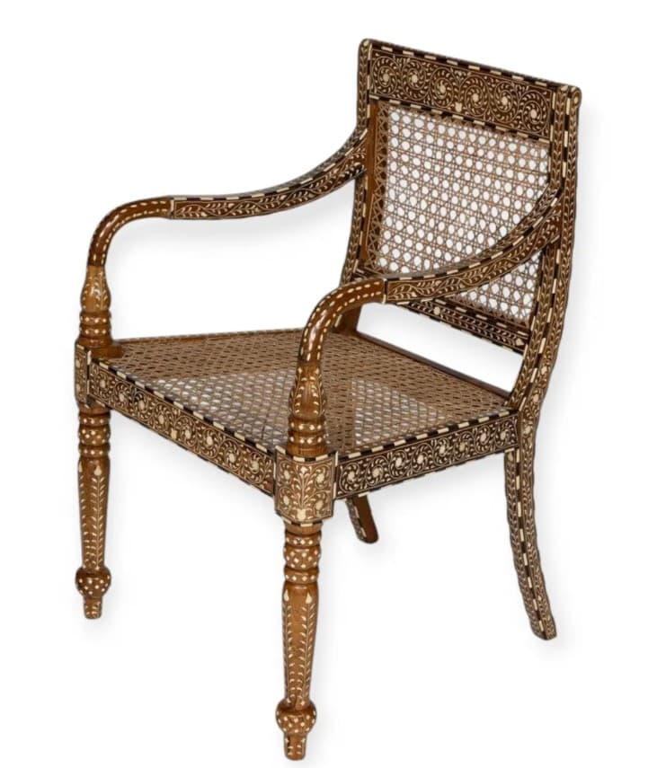 An Anglo-Indian style armchair featuring a dark-stained wooden frame extensively decorated with intricate bone or ivory inlay. It has a woven cane seat and a matching cane backrest. The arms are curved and inlaid, and the front legs are turned with ornate carvings, terminating in bun feet. The overall design is highly decorative and showcases traditional craftsmanship. — 3D model