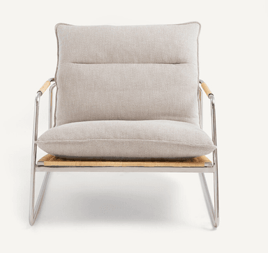 A low-profile modern lounge chair featuring a sleek, polished tubular steel frame. The seat and backrest are constructed from tightly woven natural cane, providing a textured, organic element. Thick, light-colored fabric cushions rest on the cane surfaces for comfort. The integrated armrests are part of the continuous steel frame, with a wrapped detail on the top sections. — 3D model