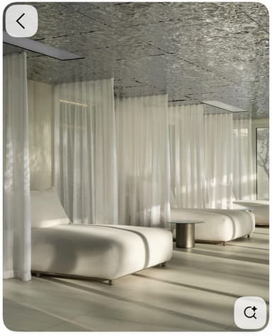 A contemporary outdoor relaxation unit featuring three individual chaise lounges arranged side-by-side, integrated within a canopy structure. The lounges are upholstered in a light-colored fabric, providing a cushioned surface. Sheer white curtains surround the perimeter, offering privacy and sun protection, while two small circular side tables are placed between the lounges for convenience. The canopy roof has a distinct textured, possibly metallic or reflective, finish. — 3D model