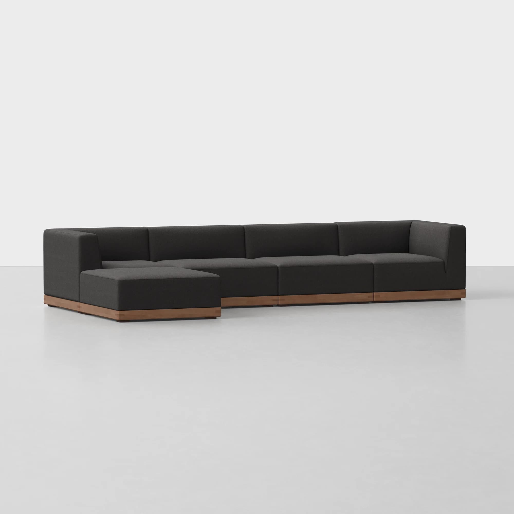 A modular sectional sofa composed of five individual units, including a corner unit and an ottoman, arranged in an L-shape. The sofa features a dark gray fabric upholstery with a low-profile, modern design. The base of each unit is accented with a continuous wooden plinth in a warm, natural wood tone. — 3D model