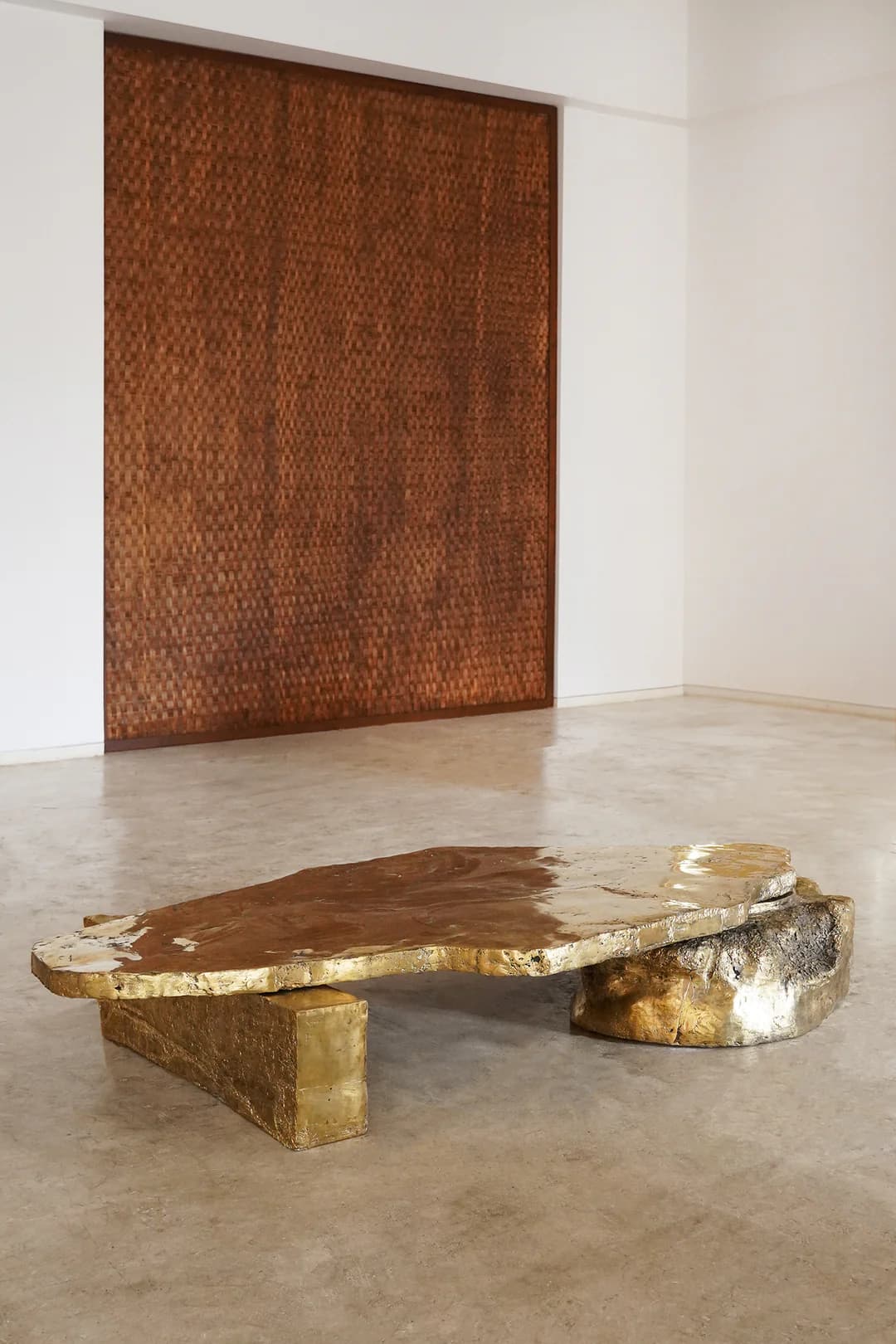 A low-profile coffee table featuring an irregularly shaped top and two distinct supports, crafted from brass and bronze. The top surface is a golden-toned, organic slab with a textured, mottled appearance. One support is a block-like form, also golden, while the other is a darker, naturalistic, rock-like bronze element. The design emphasizes raw, sculptural forms and contrasting metallic finishes. — 3D model