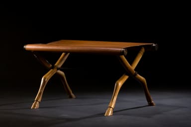 A campaign-style folding stool featuring an X-shaped wooden frame with carved details and a tan leather sling seat. The legs are notably carved to resemble animalistic or hoofed forms, adding an exotic touch. The frame is joined by visible metal hardware at the pivot points and where the leather seat attaches. — 3D model