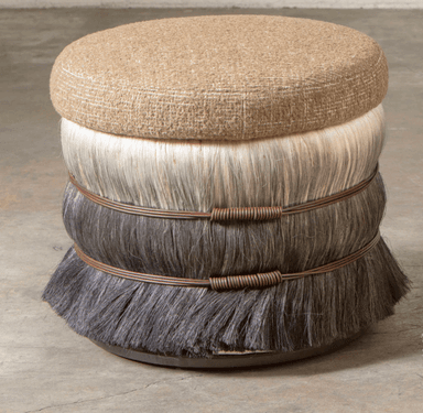 This is a unique, decorative ottoman with a round, upholstered top and a cylindrical base. The top is covered in a textured, neutral-toned fabric, possibly a linen or a blend, featuring a subtle crisscross weave. The base is constructed in tiers, with the upper tier appearing to be made of lighter colored, long hair or fiber, possibly horsehair or faux fur. The lower tier is made of darker, coarser hair or fiber, also possibly faux fur, which flares outwards towards the bottom. Two bands of thin, dark brown wire, wrapped around themselves to create a coiled effect, encircle the base between the tiers, adding an artisanal touch. The entire piece rests on a low, dark, circular base. — 3D model