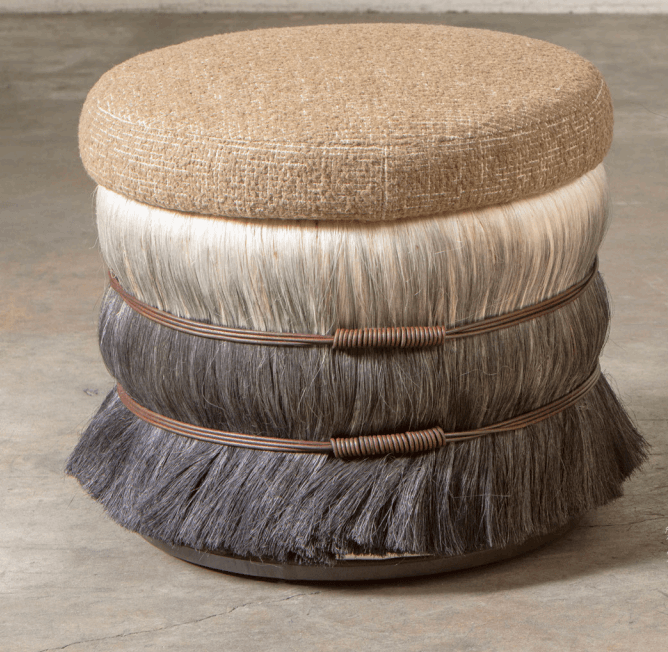 This is a unique, decorative ottoman with a round, upholstered top and a cylindrical base. The top is covered in a textured, neutral-toned fabric, possibly a linen or a blend, featuring a subtle crisscross weave. The base is constructed in tiers, with the upper tier appearing to be made of lighter colored, long hair or fiber, possibly horsehair or faux fur. The lower tier is made of darker, coarser hair or fiber, also possibly faux fur, which flares outwards towards the bottom. Two bands of thin, dark brown wire, wrapped around themselves to create a coiled effect, encircle the base between the tiers, adding an artisanal touch. The entire piece rests on a low, dark, circular base. — 3D model