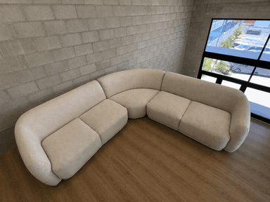 A large, modular sectional sofa forming an L-shape, featuring gently curved sections and rounded arms and backrests. It is upholstered in a light, textured fabric, giving it a contemporary and soft appearance. The sofa appears to be composed of multiple interchangeable units. — 3D model