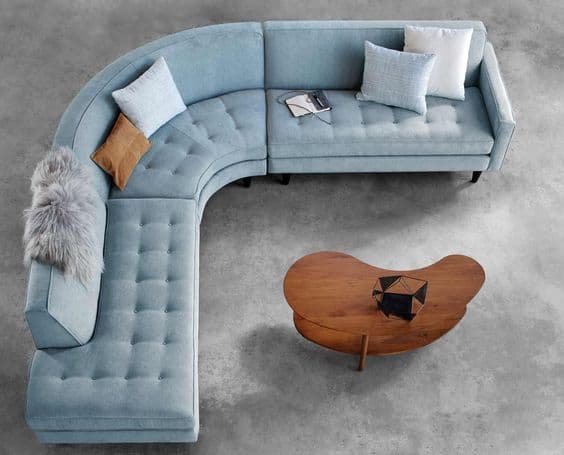 A contemporary L-shaped sectional sofa upholstered in light blue fabric, featuring a distinctive curved corner unit. The seat cushions and base of the backrest are adorned with button tufting. It includes integrated armrests at the ends and rests on dark, tapered legs. Several throw pillows in varying colors and textures are placed on the sofa. — 3D model
