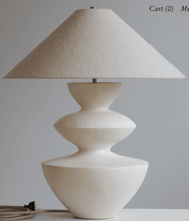A modern sculptural table lamp featuring an off-white conical fabric shade atop a similarly colored, tiered ceramic base. The base has an organic, curvilinear form, consisting of three distinct sections that narrow and widen, creating a stacked, fluid silhouette. A small brass accent is visible at the very top, connecting the shade to the lamp's body. The overall aesthetic is minimalist and contemporary, emphasizing form and texture. — 3D model