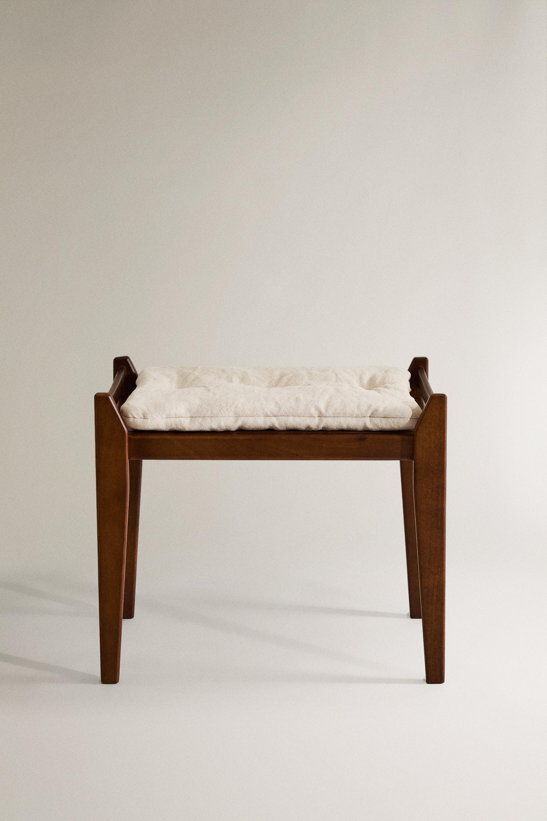 A wooden ottoman with a tufted, upholstered seat. The frame is constructed from dark wood with a visible grain. It has four tapered legs and a rectangular top with a cushioned surface covered in a light-colored, textured fabric. The seat features subtle tufting with button accents, creating a slightly padded appearance. — 3D model