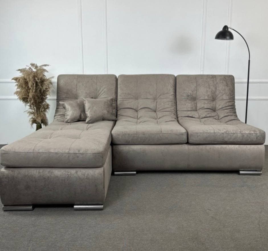 An L-shaped sectional sofa featuring a left-hand facing chaise lounge and two main seating sections. The sofa is upholstered in a light grey, textured fabric with distinctive deep tufting on the seat and back cushions. It has a low-profile design with subtly curved armrests and back, supported by small, rectangular chrome-finished feet. Two decorative throw pillows matching the upholstery are present on the chaise. — 3D model