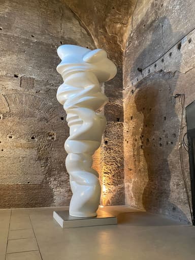 A tall, abstract floor sculpture with an organic, twisted form, rendered in a smooth, polished white material, likely resin or fiberglass. It stands on a simple square base, illuminated by uplighting, within an interior space characterized by ancient-looking brick walls. — 3D model
