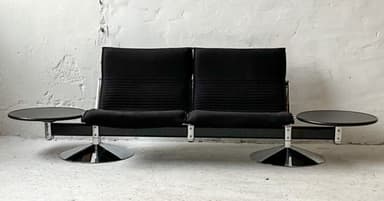 A two-seater sofa with attached side tables. The sofa features black upholstered seats and backrests with a ribbed texture on the backrests. The frame appears to be metal, possibly chrome or stainless steel, with a dark finish on the main horizontal support. Each seat has a cantilevered base with a circular, flared chrome pedestal. Attached to the horizontal frame on either side are circular, dark-colored side tables with a glossy finish, supported by metal brackets. — 3D model