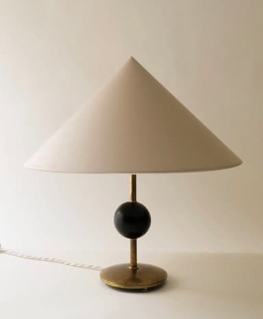 A refined table lamp featuring a wide, cream-colored conical shade. The lamp's stem connects to a dark, spherical accent, likely ebonized wood, which sits atop a cylindrical support that transitions into a circular brass base. A braided electrical cord extends from the base. — 3D model