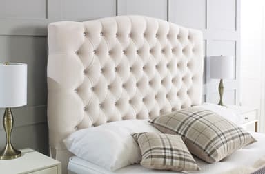 A cream-colored upholstered bed with a diamond-tufted headboard featuring crystal-like embellishments. The bed frame is also upholstered in a matching cream fabric. It is shown with a fluffy cream-colored throw blanket, two plaid throw pillows, and a gray knitted throw. The bed rests on four dark wooden carved legs. Two matching nightstands with drawers and brass accents are positioned on either side of the bed, each holding a white lampshade with a brass base. — 3D model