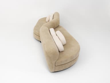 A contemporary curved modular sectional sofa upholstered in a beige, textured fabric. The sofa features a serpentine or S-shaped configuration, comprising multiple rounded segments. It is designed with a low profile and includes several rectangular, lighter-colored lumbar pillows for added comfort. The overall aesthetic is soft and organic, suitable for modern interior spaces. — 3D model