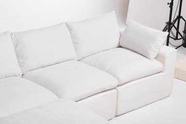 A modular sectional sofa with an attached ottoman. The sofa features deep, plush cushions and a low profile. It is upholstered in a textured white fabric. The configuration shown includes a corner section, two seat cushions, and an ottoman. The design is contemporary and emphasizes comfort. — 3D model